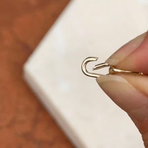 May include: Close-up of a gold-colored jewelry clasp, held against a neutral background. The clasp is open, showing the hook and the connecting piece. The metal has a shiny finish.