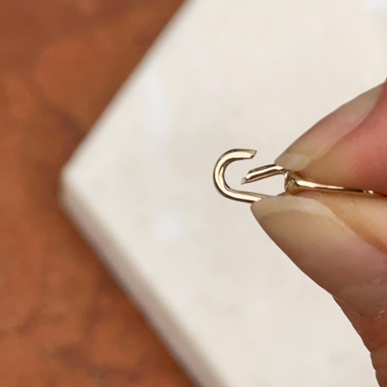 May include: Close-up of a gold-colored jewelry clasp, held against a neutral background. The clasp is open, showing the hook and the connecting piece. The metal has a shiny finish.