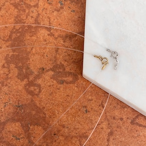 May include: Two clear elastic necklace cords with gold and silver clasps. The cords are lying on a white surface.