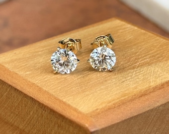May include: A pair of gold-colored stud earrings with large, round, clear gemstones. The earrings are set on a wooden surface, showcasing their sparkle and design. The earrings are a classic jewelry item.
