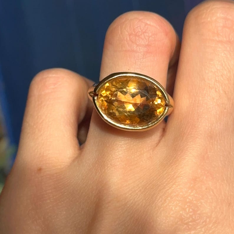 May include: A gold ring with a large, oval-shaped, yellow gemstone set in a smooth, gold setting.