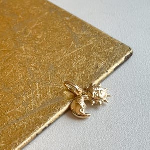 May include: A gold-colored charm featuring a sun and moon design. The sun has a smiling face with rays, and the moon has a crescent shape with a face. The charm is on a gold textured surface.