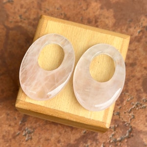 May include: Two oval-shaped pink rose quartz pendants with a hole in the center. The pendants are smooth and polished.