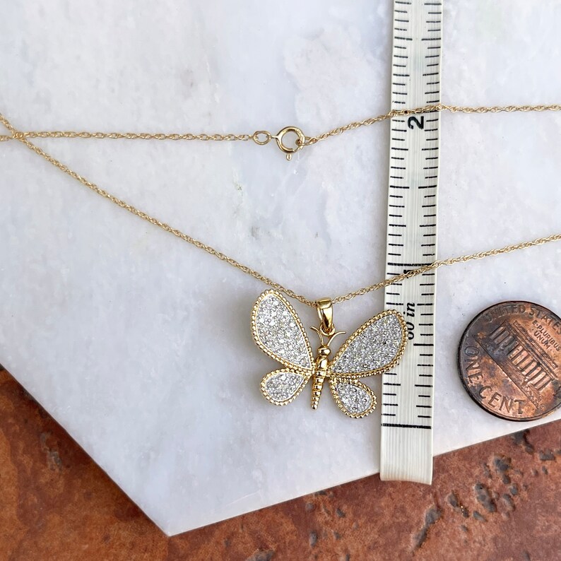 May include: A gold-colored butterfly pendant necklace with a delicate chain. The butterfly pendant is adorned with small, sparkling stones and measures approximately 1 inch in width. The necklace is displayed on a white and brown surface.
