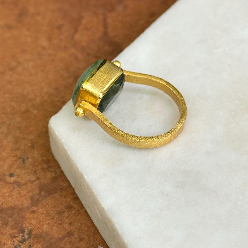 May include: A gold-colored ring with a rectangular green gemstone. The ring has a textured band and a setting that holds the gemstone securely. The ring is displayed on a white surface, showcasing its design and craftsmanship.