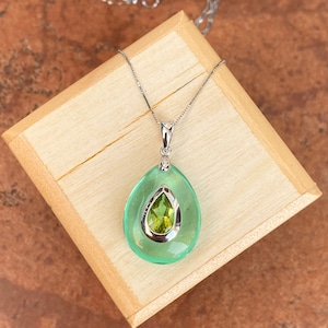 May include: A silver necklace with a pendant featuring a large, light green teardrop shape surrounding a smaller, olive-green gemstone. The pendant is set on a wooden box.
