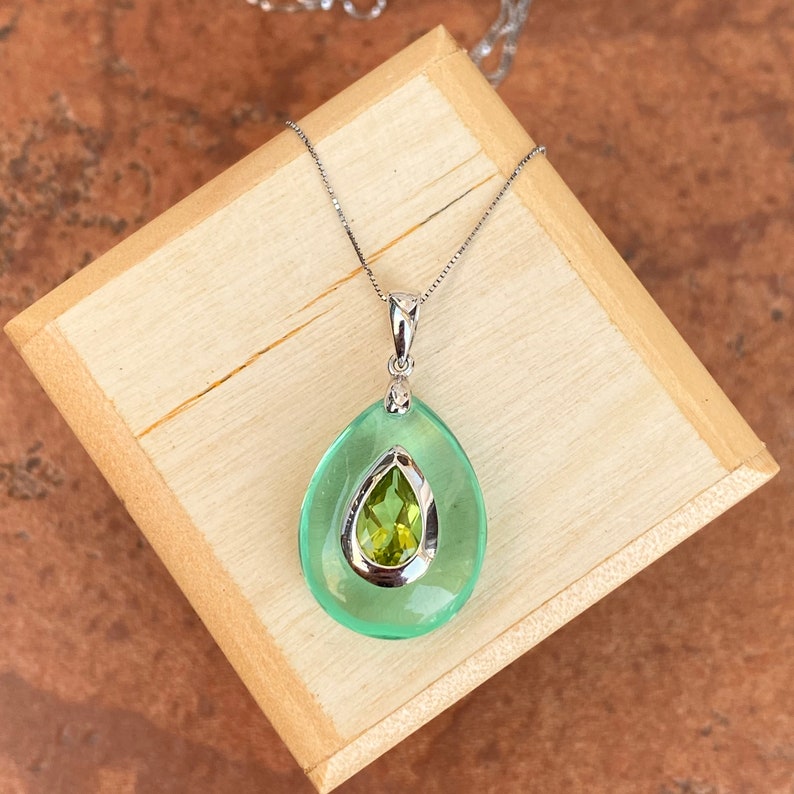 May include: A silver necklace with a pendant featuring a large, light green teardrop shape surrounding a smaller, olive-green gemstone. The pendant is set on a wooden box.