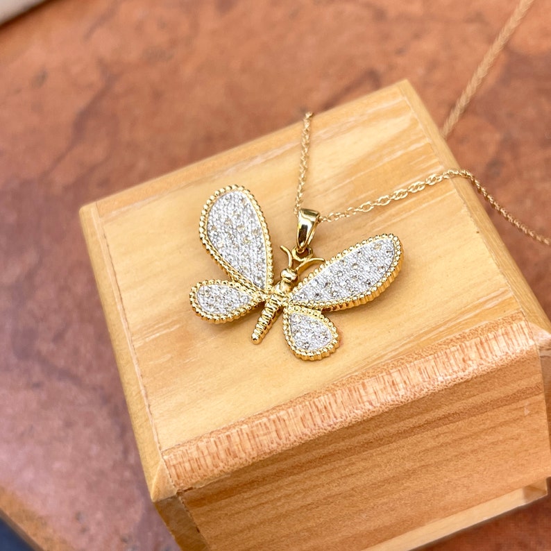 May include: A gold-colored butterfly pendant necklace with a delicate chain. The butterfly wings are encrusted with small, sparkling white stones. The necklace is displayed on a wooden surface.
