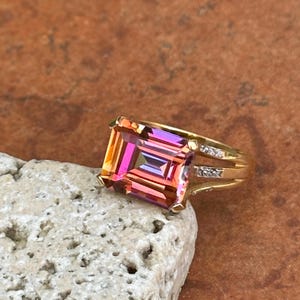 May include: A gold ring with a large, rectangular, multi-colored gemstone. The gemstone displays shades of pink, orange, and purple. The ring band has small, clear accent stones. The ring is set on a light-colored, textured stone.