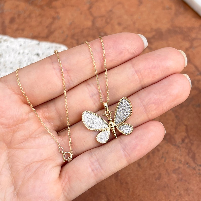 May include: A gold-colored butterfly pendant necklace. The butterfly has white, sparkling accents on its wings and a delicate gold chain. The necklace is displayed on a neutral background.