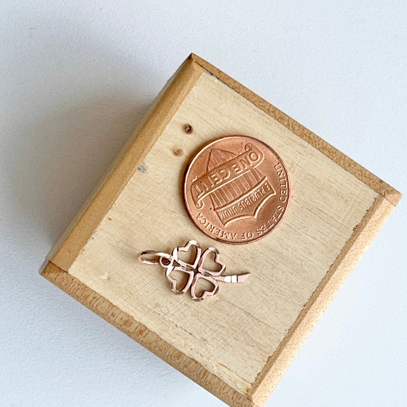 May include: A gold-toned four-leaf clover charm on a wooden surface next to a US penny.