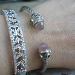 Op de afbeelding: Twee zilveren armbanden om een pols. De ene armband heeft een delicaat bloemenpatroon met heldere stenen. De andere armband heeft een gedraaid ontwerp met een roze en paarse steen.