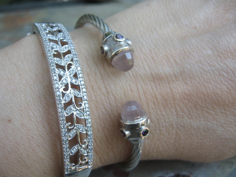 Op de afbeelding: Twee zilveren armbanden om een pols. De ene armband heeft een delicaat bloemenpatroon met heldere stenen. De andere armband heeft een gedraaid ontwerp met een roze en paarse steen.