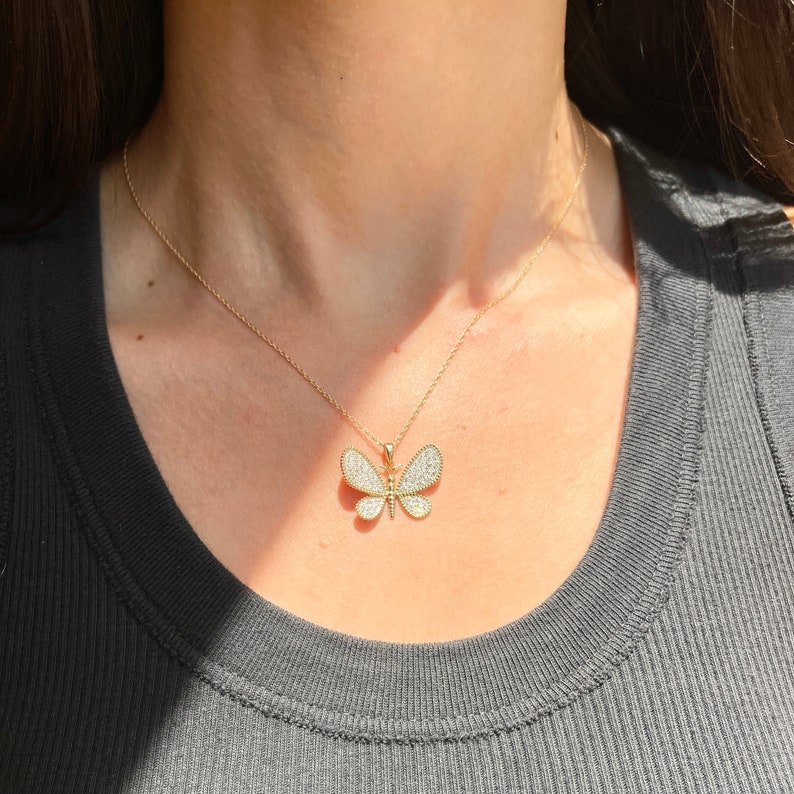 May include: A gold-colored butterfly pendant necklace. The butterfly pendant is encrusted with small, sparkling stones. The necklace is worn on a person, and the chain is delicate and gold-toned.