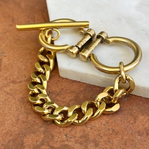 May include: A gold-colored chain bracelet with a toggle clasp. The bracelet features a thick curb chain design and a large, round clasp. The toggle clasp is a bar that fits through a loop. The bracelet is displayed on a white and brown surface.