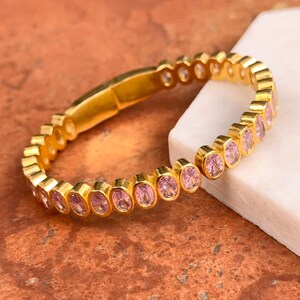 May include: A gold-colored bracelet with a series of oval, pink gemstones set in individual bezels. The bracelet is displayed on a textured, reddish-brown surface with a white, rectangular block in the background. The bracelet is a piece of jewelry.