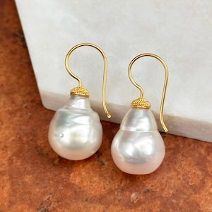 May include: A pair of pear-shaped, white pearl earrings with gold-colored hooks. The top of each pearl is capped with a textured gold design. The earrings are displayed on a white and brown surface.