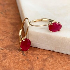 May include: A pair of gold-colored leverback earrings, each featuring a vibrant, oval-cut ruby gemstone. The earrings are set against a white and brown background, showcasing the jewelry's design and color.