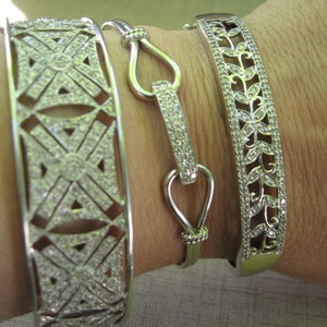 Op de afbeelding: Drie zilveren armbanden met ingewikkelde ontwerpen. De armbanden zijn versierd met kleine, heldere stenen die een sprankelend effect cre&euml;ren. De armbanden worden op de pols van een persoon gestapeld.