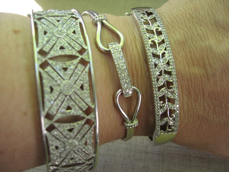 Op de afbeelding: Drie zilveren armbanden met ingewikkelde ontwerpen. De armbanden zijn versierd met kleine, heldere stenen die een sprankelend effect cre&euml;ren. De armbanden worden op de pols van een persoon gestapeld.