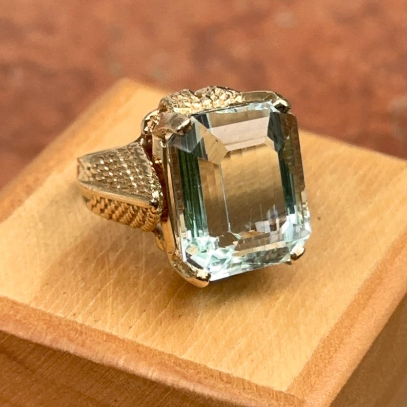 May include: A gold ring featuring a large, rectangular, light blue gemstone. The ring has intricate detailing on the band and around the gemstone setting. The ring is displayed on a wooden surface.
