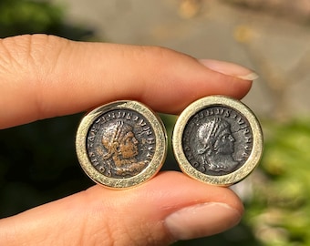 May include: Two antique coin cufflinks. Each coin features a profile of a person with a laurel wreath, set in a gold-colored bezel. The coins have a dark, aged appearance, with visible details of the faces and inscriptions. The background is blurred, suggesting an outdoor setting.
