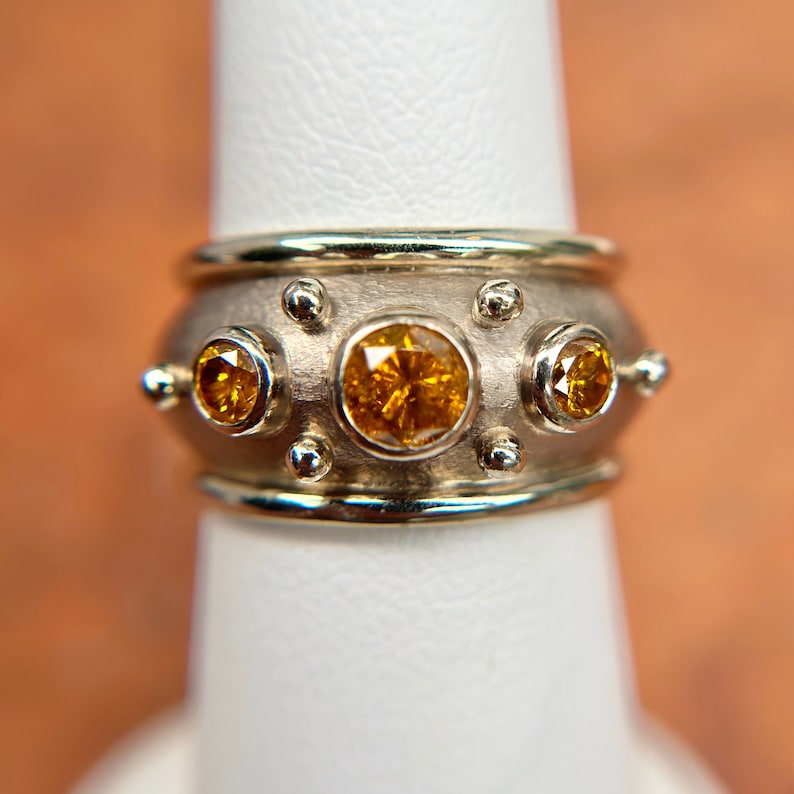 May include: A silver ring with three large, round, yellow-orange gemstones. The ring has a wide band with smaller silver accents and a gold-colored trim. The gemstones are set in a bezel setting, and the ring is displayed on a white surface.