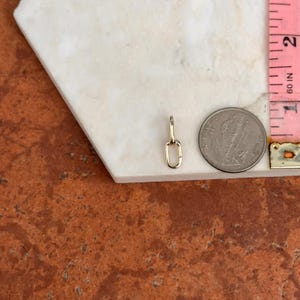 May include: A small, gold-colored chain link pendant. The pendant is next to a ruler showing measurements in inches. The pendant is on a white surface with a brown and orange background.