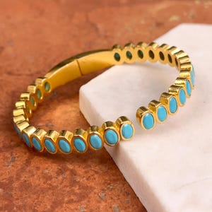 May include: A gold-colored bracelet with oval, turquoise-colored stones. The bracelet is open at one end and rests on a white surface against a brown background. The bracelet's design features a series of turquoise stones set in a gold band.