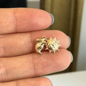May include: A gold-colored charm featuring a crescent moon and a sun with a smiling face. The sun has pointed rays around its face. The charm is small and appears to be made of metal. The charm is held between two fingers.