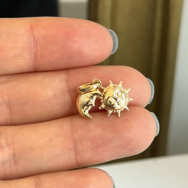 May include: A gold-colored charm featuring a crescent moon and a sun with a smiling face. The sun has pointed rays around its face. The charm is small and appears to be made of metal. The charm is held between two fingers.