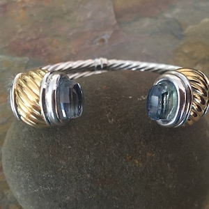 May include: A silver and gold-tone cuff bracelet with blue gemstone accents. The bracelet features a twisted cable design and gold-tone end caps with a ribbed texture. The gemstones are a light blue color.