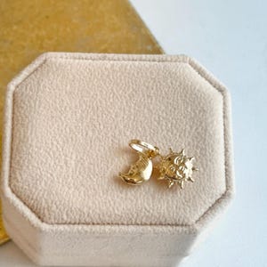 May include: A gold-colored pendant featuring a crescent moon and a sun with rays. The celestial charms are displayed on a beige velvet surface, suggesting a jewelry or accessory item. The pendant is likely for a necklace or bracelet.