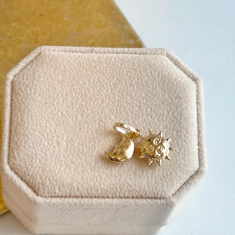 May include: A gold-colored pendant featuring a crescent moon and a sun with rays. The celestial charms are displayed on a beige velvet surface, suggesting a jewelry or accessory item. The pendant is likely for a necklace or bracelet.