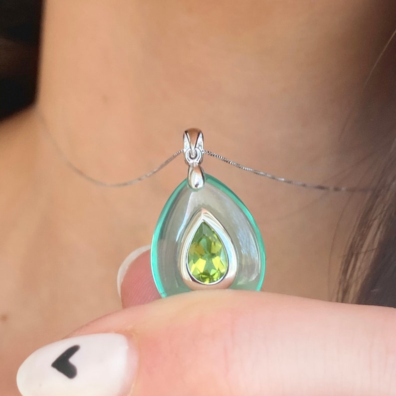 May include: A silver pendant necklace featuring a teardrop-shaped, light green gemstone. The pendant has a clear, outer layer and a smaller, pear-shaped, lime green stone at its center. The necklace is displayed against a blurred background.