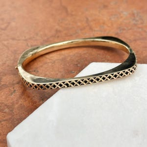 Könnte beinhalten: Ein goldenes Armreif-Armband mit einem schwarzen geometrischen Muster. Das Armband ist quadratisch und hat einen Scharnierverschluss.