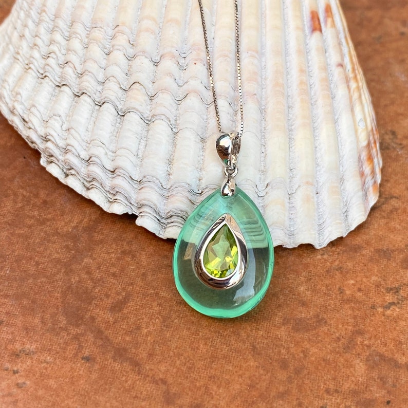 May include: A silver necklace with a teardrop-shaped pendant. The pendant has a light green outer layer and a smaller, pear-shaped, light green stone in the center. The necklace is displayed on a white seashell.
