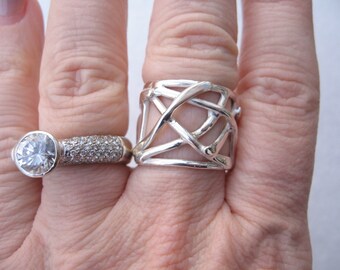 May include: Two silver rings on a hand. The ring on the index finger is a wide band with a geometric design. The ring on the middle finger is a thin band with a large, clear stone and smaller stones around it.