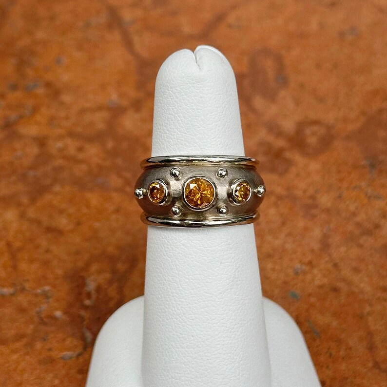 May include: A silver-toned ring with three large and four small round, yellow gemstones. The ring has a wide band with a textured surface and small, raised dots. The ring is displayed on a white finger.