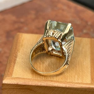 May include: A gold ring featuring a large, rectangular, light blue gemstone. The ring has intricate detailing on the band and around the gemstone setting. The ring is displayed on a wooden surface.