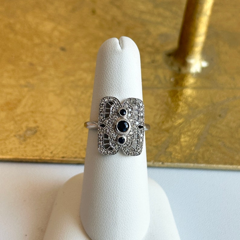 May include: A silver ring with a black stone centre and a filigree design. The ring is on a white display stand.