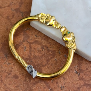 May include: A gold-tone metal bangle bracelet featuring two sculpted horse head terminals facing each other. One horse head has a small clear stone eye, and the other has a small green stone eye. The band includes a textured ribbed section near the clasp.