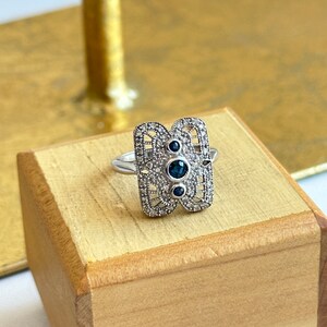 May include: A silver ring with a rectangular design featuring a filigree pattern and three blue gemstones.