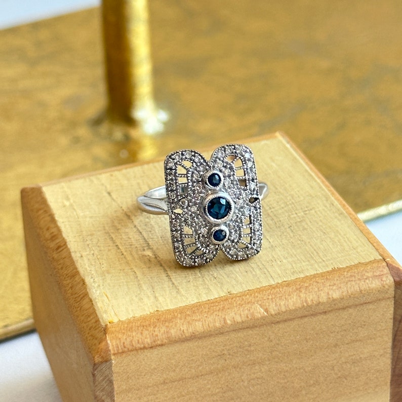 May include: A silver ring with a rectangular design featuring a filigree pattern and three blue gemstones.