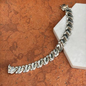 May include: A silver-toned bracelet with a series of stylized, leaf-shaped links. The bracelet is laid out on a white surface, with a clasp at one end. The links are smooth and reflective, catching the light.