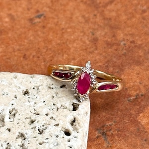 May include: A gold ring featuring a central marquise-cut red ruby surrounded by small clear stones. The band includes two additional red rubies set into the gold on either side of the centre stone, creating a symmetrical and detailed design.