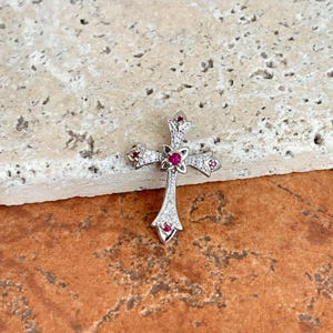 May include: A silver-colored cross pendant with a central ruby and smaller rubies at the ends of the cross arms. The cross is adorned with small, clear gemstones. The pendant is displayed on a textured, light-colored surface.