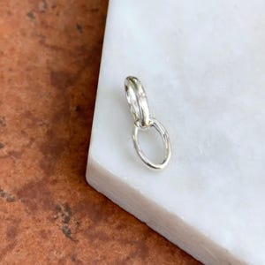 May include: A small, silver-colored pendant with an oval loop connected to a smaller, rounded loop. The pendant is displayed on a white surface with a brown and orange background.