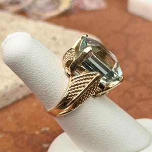 May include: A gold ring featuring a large, rectangular, light blue gemstone. The ring has an ornate, textured gold band and setting, with intricate detailing around the gemstone. The ring is displayed on a white finger form.