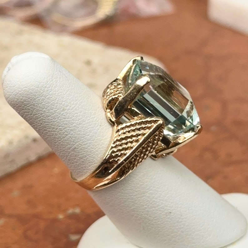 May include: A gold ring featuring a large, rectangular, light blue gemstone. The ring has an ornate, textured gold band and setting, with intricate detailing around the gemstone. The ring is displayed on a white finger form.